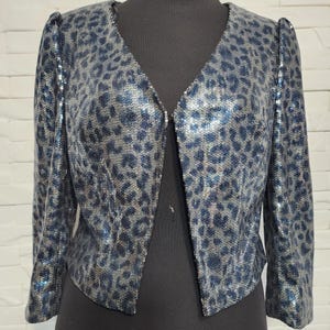Könnte beinhalten: Eine Paillettenjacke mit Leopardenmuster in Blau- und Silbertönen. Die Jacke hat einen V-Ausschnitt und Dreiviertelärmel. Das Kleidungsstück wird auf einer schwarzen Schaufensterpuppe präsentiert.