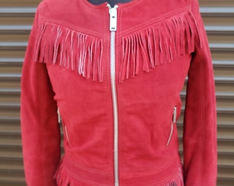 Puede incluir: Chaqueta de ante roja con flecos en el pecho y el dobladillo. La chaqueta tiene cierre de cremallera plateada y dos pequeños bolsillos con cremallera. Se muestra en un maniquí negro.