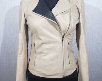 Puede incluir: Chaqueta de cuero beige con forro interior negro y cremalleras plateadas. La chaqueta tiene un cierre de cremallera asimétrico clásico, dos bolsillos delanteros y mangas largas. Se muestra en un maniquí negro.
