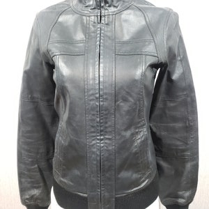 Pode incluir: Casaco de couro preto com fecho de correr, gola alta e punhos e bainha canelados. O casaco tem um estilo bomber clássico com detalhes costurados nos ombros e no peito.