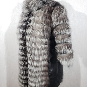K&ouml;nnte beinhalten: Ein luxuri&ouml;ser Silberfuchsfellmantel mit schwarzem Lederbesatz. Der Mantel hat lange &Auml;rmel und einen hohen Kragen. Das Fell ist eine Mischung aus Silber, Grau und Schwarz und erzeugt eine auff&auml;llige visuelle Textur.