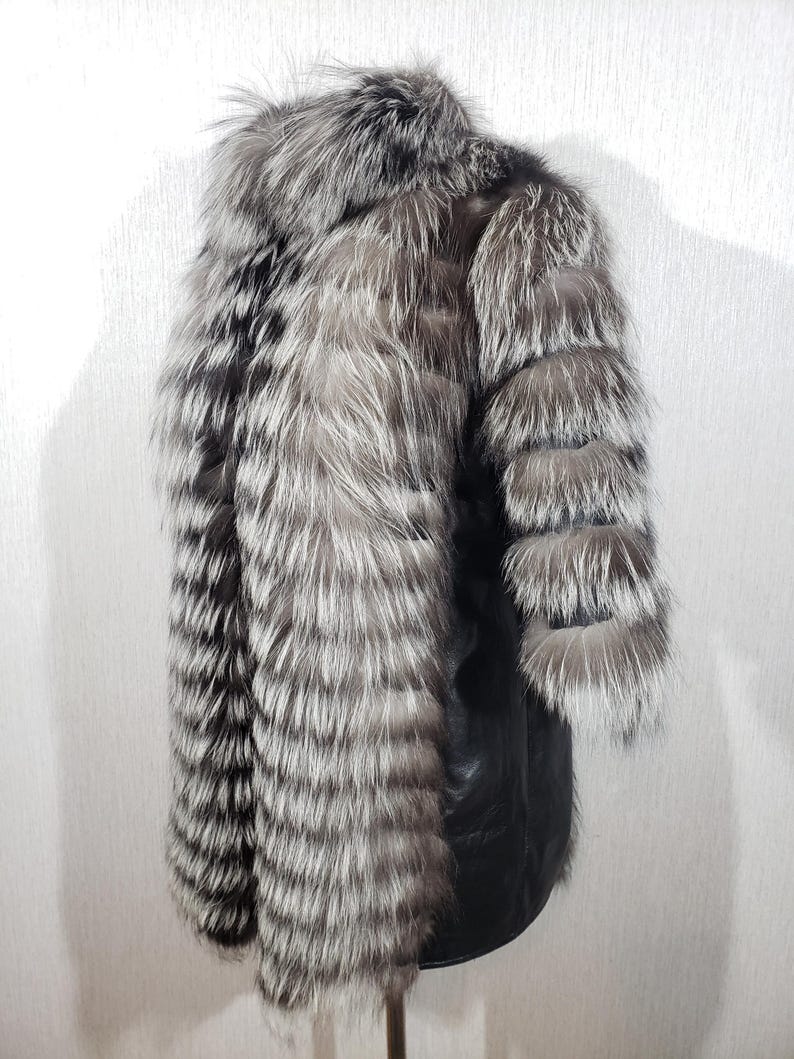 K&ouml;nnte beinhalten: Ein luxuri&ouml;ser Silberfuchsfellmantel mit schwarzem Lederbesatz. Der Mantel hat lange &Auml;rmel und einen hohen Kragen. Das Fell ist eine Mischung aus Silber, Grau und Schwarz und erzeugt eine auff&auml;llige visuelle Textur.