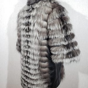 K&ouml;nnte beinhalten: Ein silber-grauer Pelzmantel mit schwarzem Lederboden. Der Mantel hat einen gro&szlig;en Kragen und lange &Auml;rmel. Das Fell ist in horizontalen Streifen angeordnet, wodurch ein strukturiertes Aussehen entsteht. Der Mantel wird auf einem St&auml;nder pr&auml;sentiert.