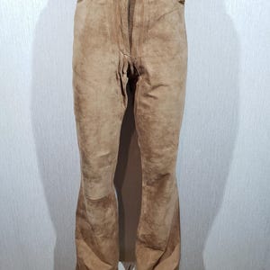 May include: Tan suede trousers with a button and zip fastening. The trousers feature a classic five-pocket design and a slight flare at the hem. The trousers are displayed on a mannequin.