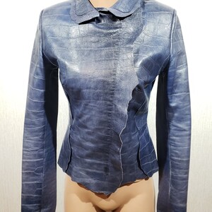 May include: A blue leather jacket with a textured, crocodile-like pattern. The jacket has a unique collar and a fitted silhouette. The color is a muted blue, and the jacket appears to be made of high-quality leather.