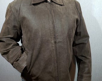 Può includere: Giacca in pelle marrone con chiusura a zip e collo. La giacca ha due tasche anteriori e un aspetto leggermente usurato. Le maniche hanno un semplice design a polsino. La giacca è indossata da una persona.