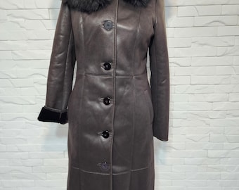 Può includere: Lungo cappotto in pelle marrone con cappuccio e polsini foderati in pelliccia nera. Il cappotto presenta una chiusura frontale con bottoni e una vestibilità su misura. Il cappotto è esposto contro un muro di mattoni bianchi.