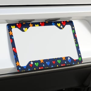 May include: A rectangular license plate frame featuring a dark blue background with a repeating pixel art pattern of red, green, yellow, and blue hearts and stars. The frame has four mounting holes and is attached to a white vehicle bumper.