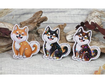 May include: Three holographic stickers of Shiba Inu dogs in different colors: orange, black, and brown. The dogs are sitting with their tongues sticking out and have a sparkly, iridescent finish.