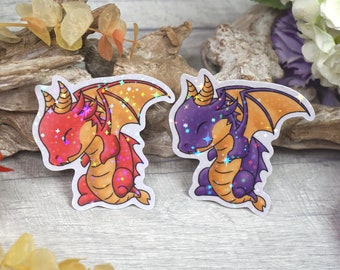 Puede incluir: Dos pegatinas holográficas de dragones de dibujos animados con los ojos cerrados. El dragón de la izquierda es rojo con alas naranjas y el dragón de la derecha es morado con alas naranjas.