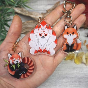 Könnte beinhalten: Drei Schlüsselanhänger aus klarem Acryl mit Cartoon-Füchsen und einem Roten Panda. Die Schlüsselanhänger haben goldfarbene Verschlüsse. Ein Schlüsselanhänger zeigt einen Roten Panda, der Bambus frisst. Ein weiterer zeigt einen neunschwänzigen Fuchs. Der dritte Schlüsselanhänger ist ein Fuchs.