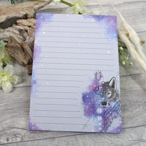 May include: A lined notepad with a watercolor galaxy design and a wolf's head illustration. The wolf's head is surrounded by a geometric design with circles and lines.