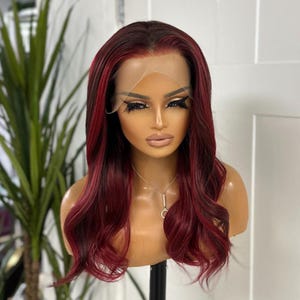 May include: A burgundy and black ombre wig with long, wavy hair. The wig is styled on a mannequin head with a natural-looking hairline. The wig has a lace front and is designed to look like natural hair.