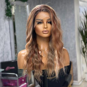 May include: A wavy, long wig with a lace front, displayed on a mannequin head. The wig features a blend of brown, blonde, and reddish-brown tones. The hair is styled with loose waves, and the mannequin has makeup and a necklace.