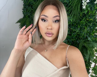 Puede incluir: Una mujer con el pelo rubio claro lleva un top beige de un solo hombro. Está de pie frente a una pared verde con flores blancas.