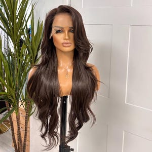 Puede incluir: Una peluca marrón con cabello largo y ondulado se muestra en una cabeza de maniquí. La peluca presenta un frente de encaje y está peinada con una raya a un lado. El maniquí está sobre un soporte negro, con una planta visible en el fondo.