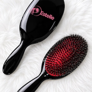 May include: Two oval hairbrushes with glossy black handles and heads. One brush shows the back with a pink logo reading Estelle and a stylised profile graphic. The other brush shows the front with black bristles set into a vibrant red cushion.