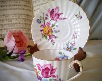 May include: A white teacup and saucer with a floral pattern. The cup and saucer have a gold trim and feature pink, yellow, and blue flowers.