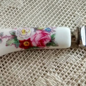May include: A white ceramic butter knife with a floral design and a silver blade. The blade is engraved with "Royal Crown Derby" and "England".