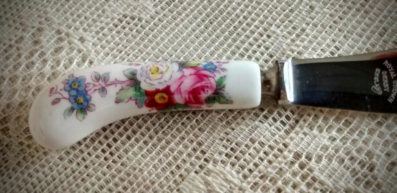 May include: A white ceramic butter knife with a floral design and a silver blade. The blade is engraved with "Royal Crown Derby" and "England".