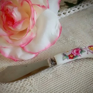 May include: A vintage butter knife with a white porcelain handle featuring a floral design in shades of pink, red, yellow, and green. The blade is stainless steel.