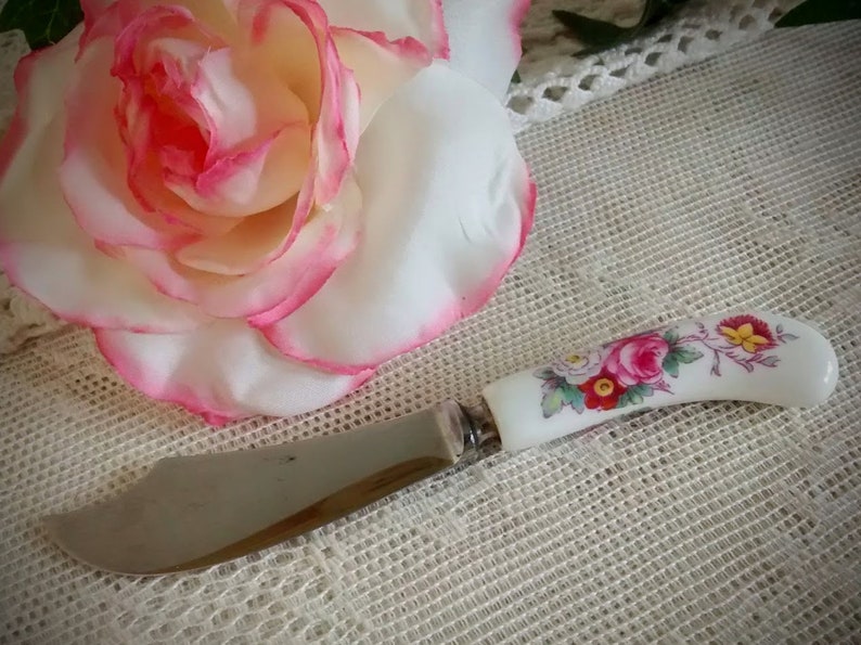 May include: A vintage butter knife with a white porcelain handle featuring a floral design in shades of pink, red, yellow, and green. The blade is stainless steel.