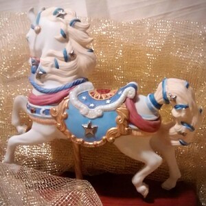 May include: A white porcelain carousel horse figurine with blue, red, and gold accents. The horse has a flowing mane and tail with stars and a blue and gold saddle.