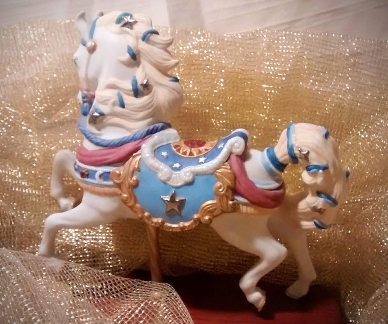 May include: A white porcelain carousel horse figurine with blue, red, and gold accents. The horse has a flowing mane and tail with stars and a blue and gold saddle.