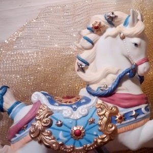 May include: A white porcelain carousel horse with blue, gold, and red details. The horse has a flowing mane and tail, and is decorated with stars and other festive designs.