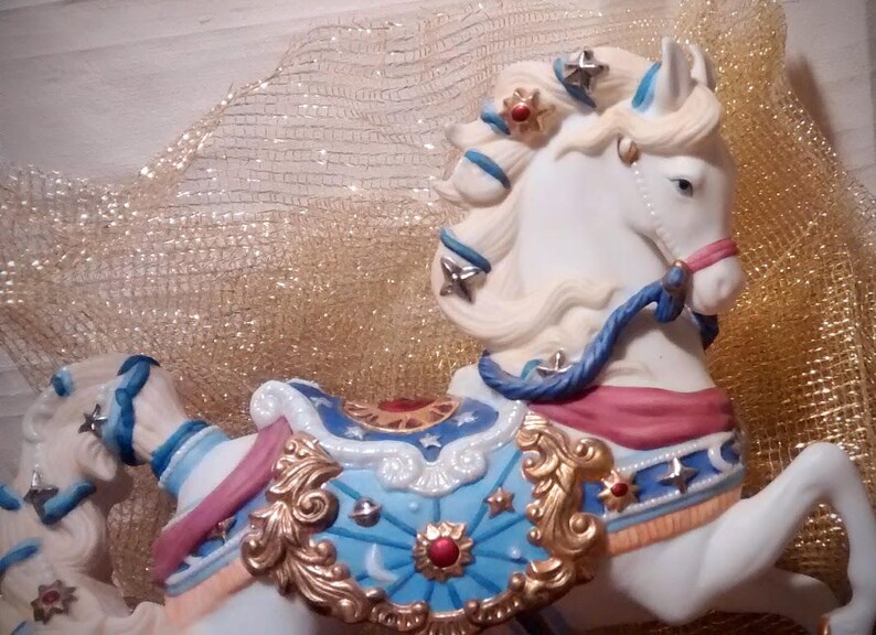 May include: A white porcelain carousel horse with blue, gold, and red details. The horse has a flowing mane and tail, and is decorated with stars and other festive designs.