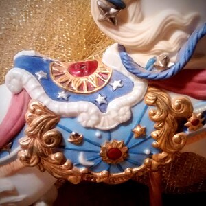May include: A white porcelain carousel horse with a blue, gold, and red saddle. The saddle is decorated with stars, moons, and suns.