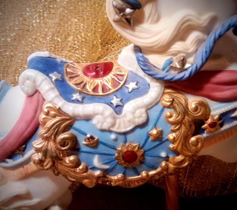 May include: A white porcelain carousel horse with a blue, gold, and red saddle. The saddle is decorated with stars, moons, and suns.