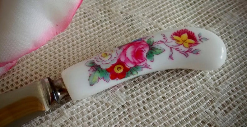 May include: A white ceramic butter knife with a floral design. The handle features pink, red, white, and yellow flowers with green leaves.