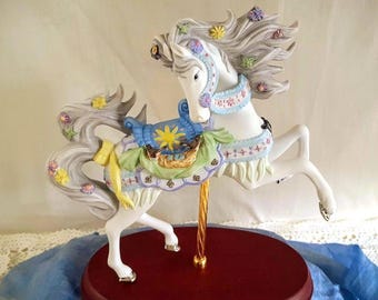 Può includere: Una statuetta di cavallo da giostra bianca con criniera e coda grigie fluenti, adornata da fiori colorati. Il cavallo ha una sella blu e verde con un disegno a stella gialla e un palo dorato. La base è di colore rosso scuro.