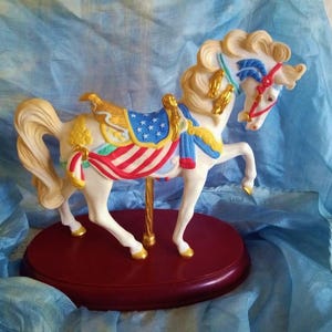 May include: A white porcelain carousel horse figurine with a red, white, and blue saddle and a gold-painted mane and tail. The horse is standing on a red wooden base.