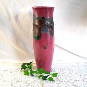 May include: A tall, cylindrical vase in shades of pink and burgundy. The upper portion features a textured, dark brown and gray design with green accents. The vase is set against a white background with a sprig of green leaves.