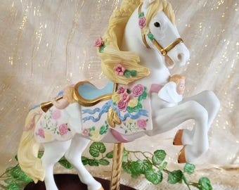 Può includere: Statuetta di cavallo da giostra bianca con criniera e coda gialle fluenti, adornata con rose rosa e gialle. Il cavallo ha una sella azzurra e un palo color oro, montato su una base marrone scuro.