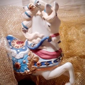 May include: White porcelain carousel horse figurine with blue, red, gold, and white painted details. The horse has a flowing mane and tail and is decorated with stars and moons.