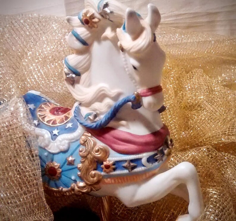 May include: White porcelain carousel horse figurine with blue, red, gold, and white painted details. The horse has a flowing mane and tail and is decorated with stars and moons.