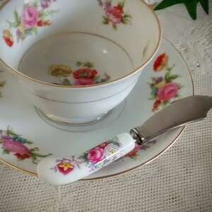 May include: A white porcelain teacup and saucer with a floral pattern. The saucer has a gold trim. A small, white butter knife with a floral pattern on the handle is resting on the saucer.