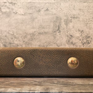May include: A brown leather box with two round, inset images of people riding horses. The box has a textured surface.