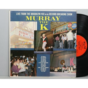 May include: A vintage vinyl record album cover featuring a photo of Murray the K, a popular radio DJ, performing live at the Brooklyn Fox Theater. The cover features a collage of photos of Murray the K on stage, with a list of artists who appeared on his show, including The Angels, The Chiffons, The Dovells, The Drifters, Jan and Dean, Jay and the Americans, Ben E. King, The Miracles, Gene Pitney, Randy and the Rainbows, The Ronettes, The Shirelles, and The Tymes.
