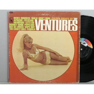 May include: A vintage vinyl record album cover featuring a woman in a gold bikini lying on a white background. The album cover is orange and features the text "Golden Greats by The Ventures" and a list of songs including "Rebel Rouser", "Walk, Don't Run", "Apache", "Telstar", "Memphis", "Let's Go", "Honky Tonk", "Pipeline", "Out of Limits", "Wipe-Out", "The Lonely Bull", and "Tequila".