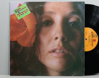 May include: Album cover featuring Maria Muldaur with a large orange flower in her hair. The album title, "Waitress in a Donut Shop," is in a green and white box. A black vinyl record is visible on the right side of the image.