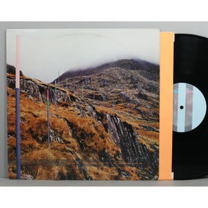 May include: A vinyl record album featuring a landscape image of a mountain range with colorful vertical lines. The album cover has a muted color palette with a black vinyl record visible on the right side.