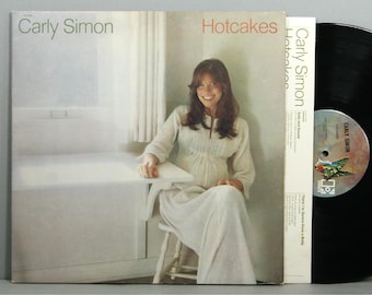 May include: Carly Simon's "Hotcakes" album cover. The album cover features a woman in a white dress sitting at a table. The album title is in red text. A black vinyl record is visible on the right.