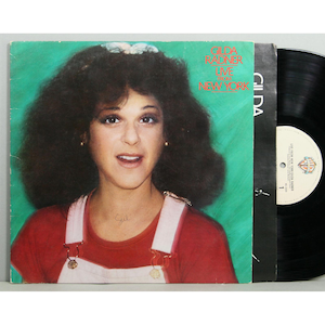 May include: Album cover for Gilda Radner's "Live From New York" record. The cover features a portrait of Gilda Radner in a red top and white overalls against a green background. A black vinyl record is visible to the right.