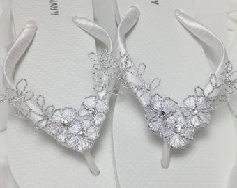 May include: White flip flops with silver floral embellishments. The flowers have a delicate lace design and are accented with small rhinestones.