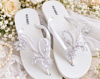 May include: A pair of white Old Navy flip-flops featuring satin straps adorned with silver floral embroidery, white pearls, and small flower appliqués. The footbeds are textured white rubber with the Old Navy brand name printed in black.