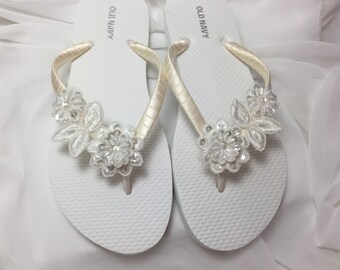 May include: White flip-flops with ivory satin straps and floral embellishments. The flowers are made of white lace and adorned with clear sequins and beads. The flip-flops are a summer accessory.